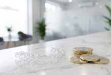 A photorealistic close-up of a clear Invisalign aligner next to a stack of blurred British Pound coins on a white marble surface, symbolizing the cost of orthodontic treatment in the UK.