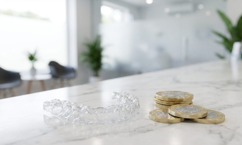 A photorealistic close-up of a clear Invisalign aligner next to a stack of blurred British Pound coins on a white marble surface, symbolizing the cost of orthodontic treatment in the UK.