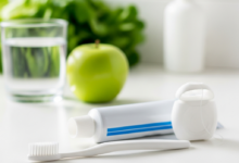 Close-up of a new soft-bristled toothbrush, an unbranded tube of toothpaste, and dental floss on a clean, bright surface. In the blurred background, there's a clear glass of water and healthy food items like an apple and leafy greens, symbolizing a comprehensive approach to oral health. The scene is brightly lit, emphasizing cleanliness and well-being.