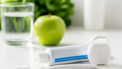Close-up of a new soft-bristled toothbrush, an unbranded tube of toothpaste, and dental floss on a clean, bright surface. In the blurred background, there's a clear glass of water and healthy food items like an apple and leafy greens, symbolizing a comprehensive approach to oral health. The scene is brightly lit, emphasizing cleanliness and well-being.