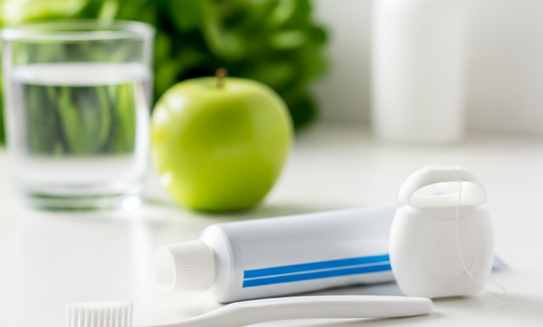 Close-up of a new soft-bristled toothbrush, an unbranded tube of toothpaste, and dental floss on a clean, bright surface. In the blurred background, there's a clear glass of water and healthy food items like an apple and leafy greens, symbolizing a comprehensive approach to oral health. The scene is brightly lit, emphasizing cleanliness and well-being.