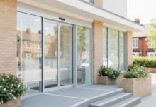 A modern, clean dental clinic building entrance with light-colored walls, large windows, and an inviting doorway, bathed in natural daylight. The background is softly blurred, hinting at a typical British street or residential area. There is no visible text, logos, or branding.